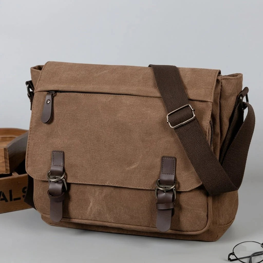 A brown canvas messenger bag with dark brown leather straps and metal buckles, sitting on a grey surface.
