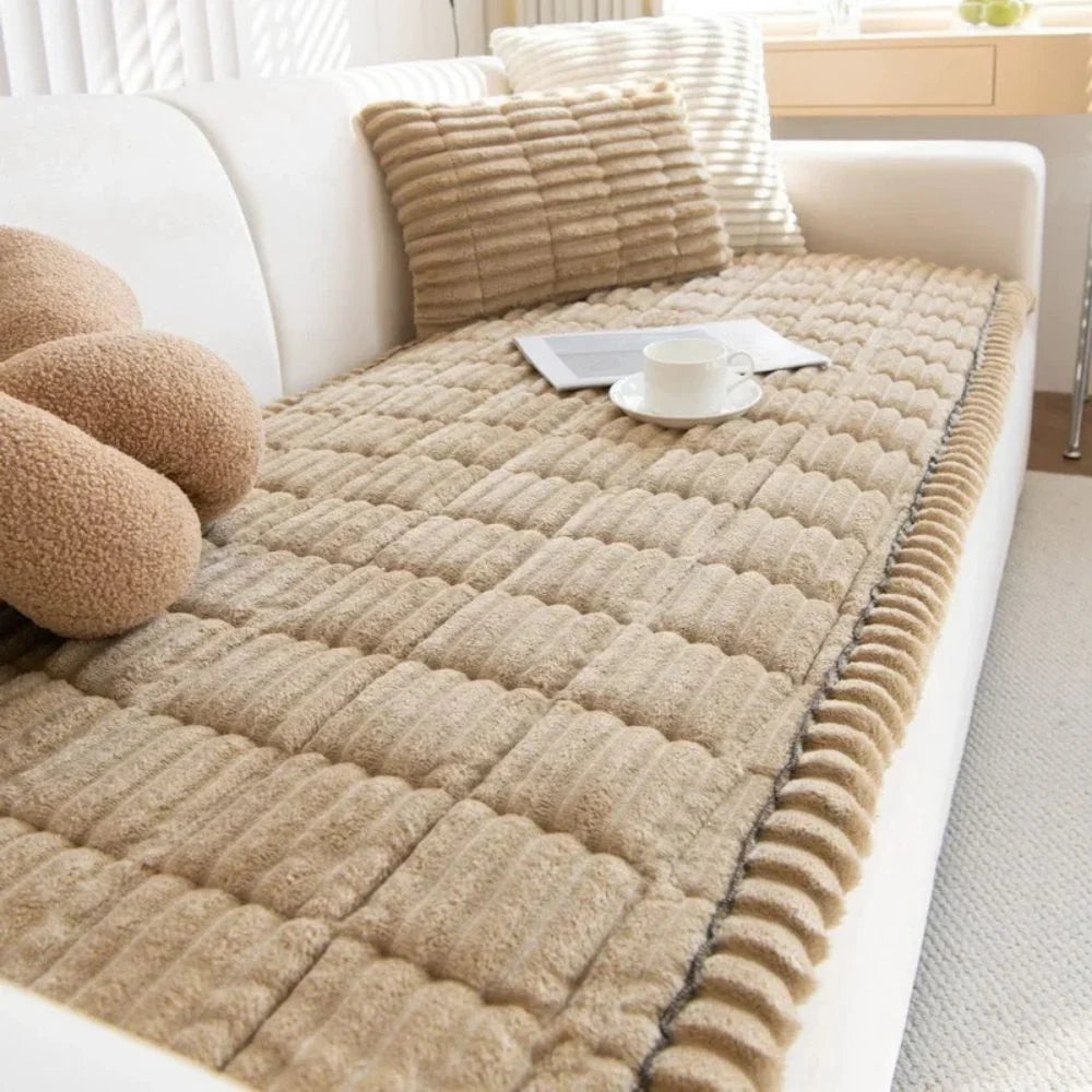 A beige, quilted, non-slip sofa cover draped over a white couch with matching plush cushions and a coffee cup on top.