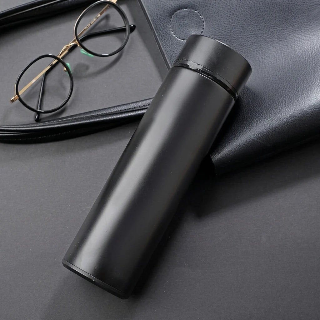 A sleek black insulated stainless steel water bottle with a digital LED display lid, placed next to round glasses.