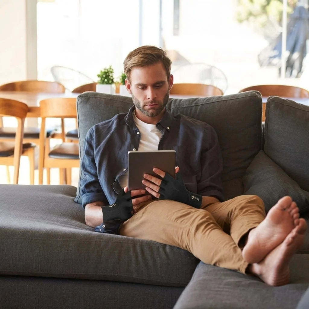 A man sitting on a grey sofa using a tablet while wearing black compression gloves with an open-finger design.