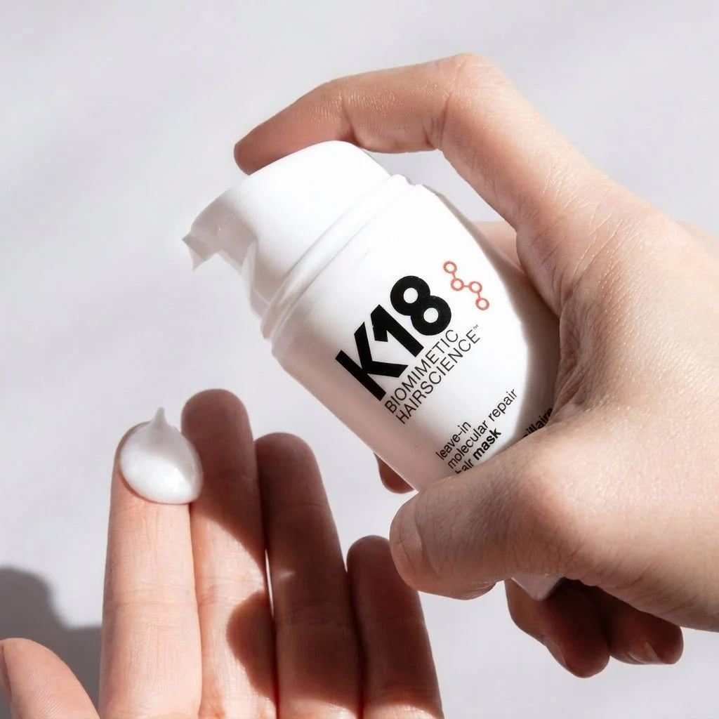 A hand dispensing a small amount of white K18 leave-in molecular repair hair mask cream onto a palm.