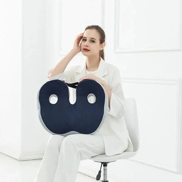 A woman in a white suit sitting on a chair and holding a dark blue ergonomic memory foam seat cushion.