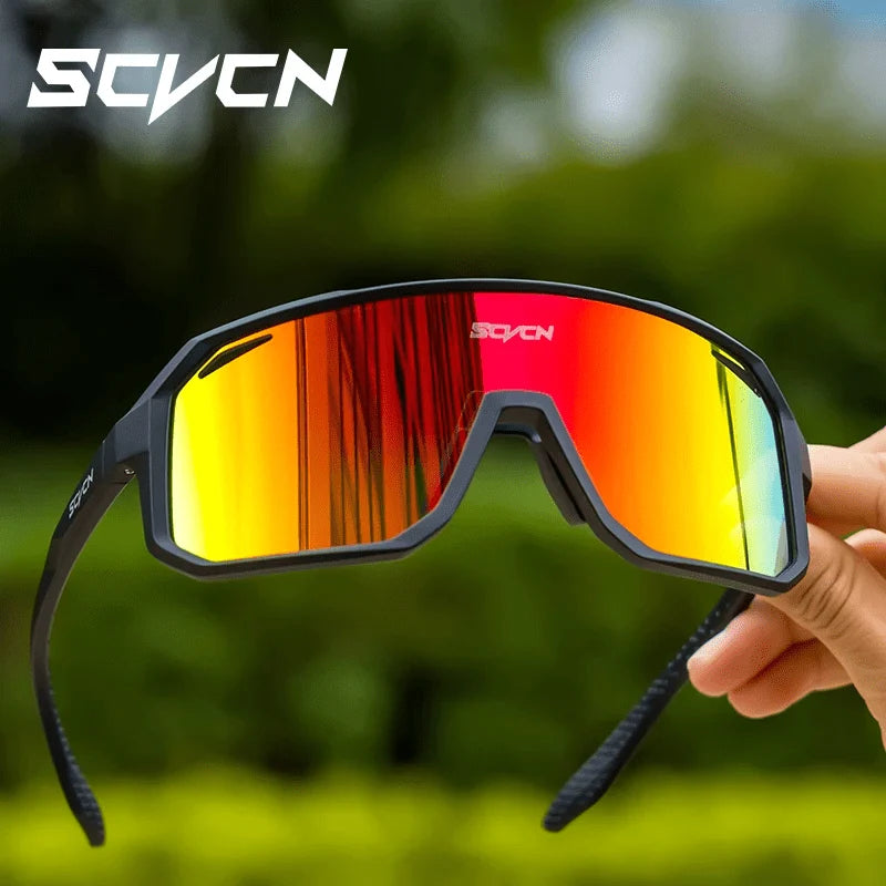 A hand holds a pair of black cycling sunglasses with vibrant red and orange reflective lenses against a blurred green background.