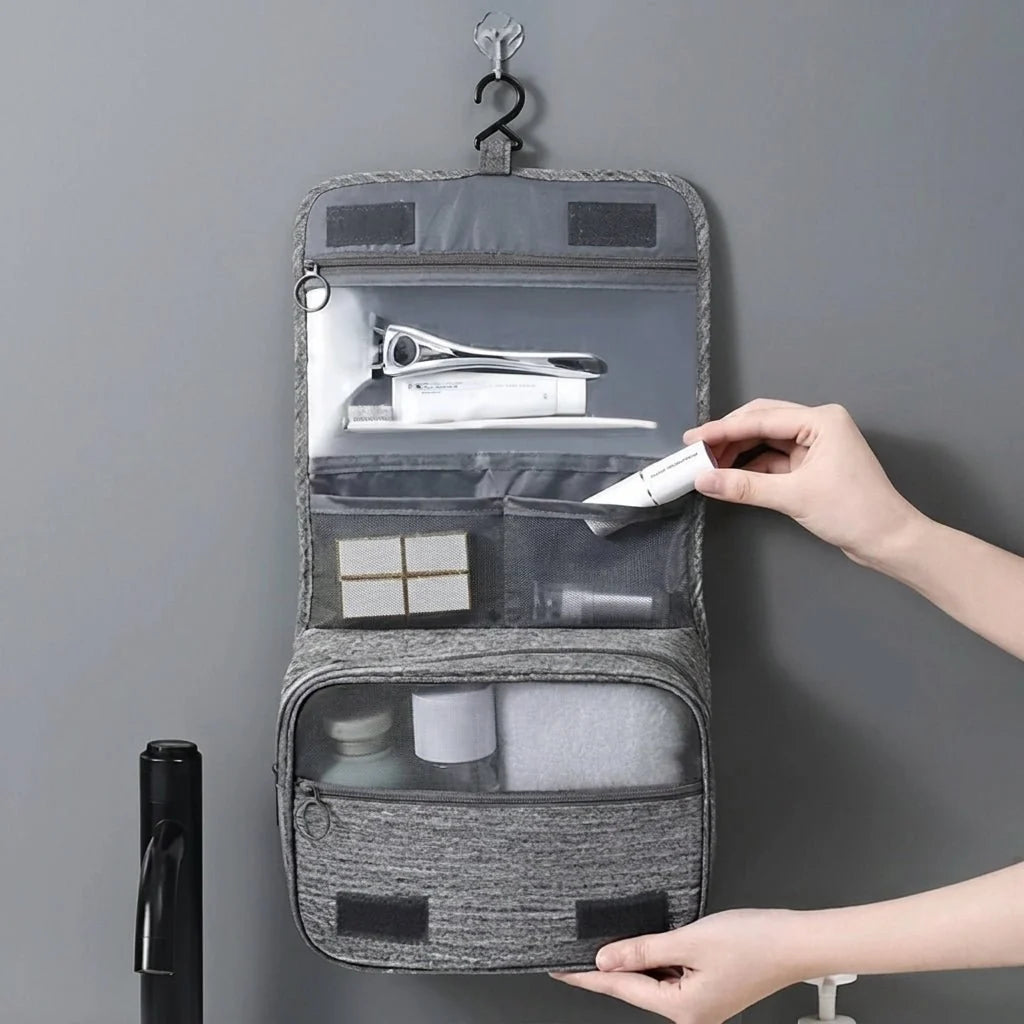 A person holding a grey hanging toiletry bag filled with travel-sized grooming items against a plain wall.