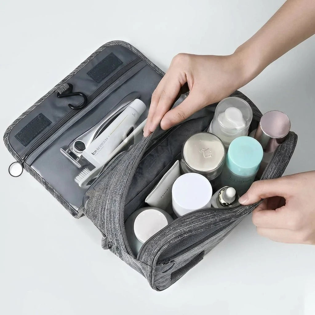 A pair of hands opening a grey, hanging toiletry bag filled with various skincare bottles and grooming items.