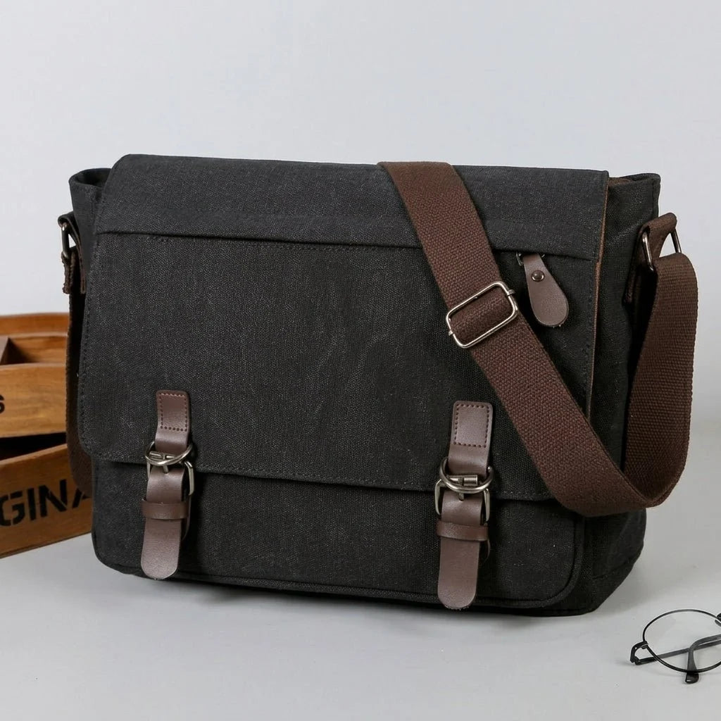 A black canvas messenger bag with brown leather straps and buckles, sitting on a grey surface next to a pair of glasses.
