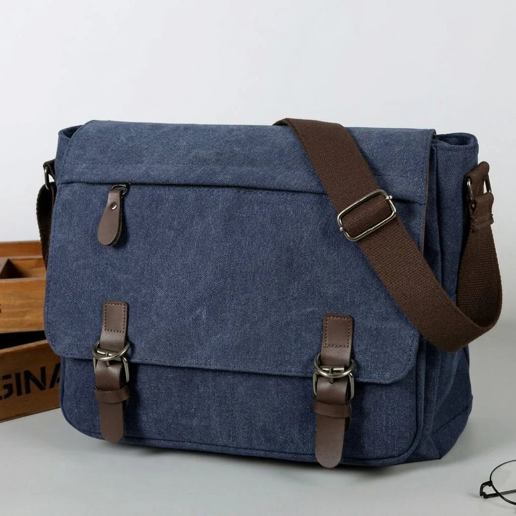 A blue canvas messenger bag with brown leather-look straps and buckles, sitting next to a wooden crate.