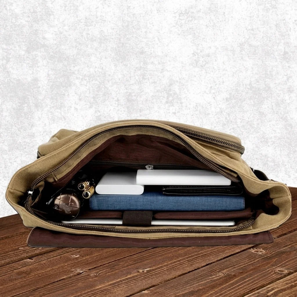An open tan canvas messenger bag sitting on a wooden table, containing a laptop, a blue notebook, and a power bank.