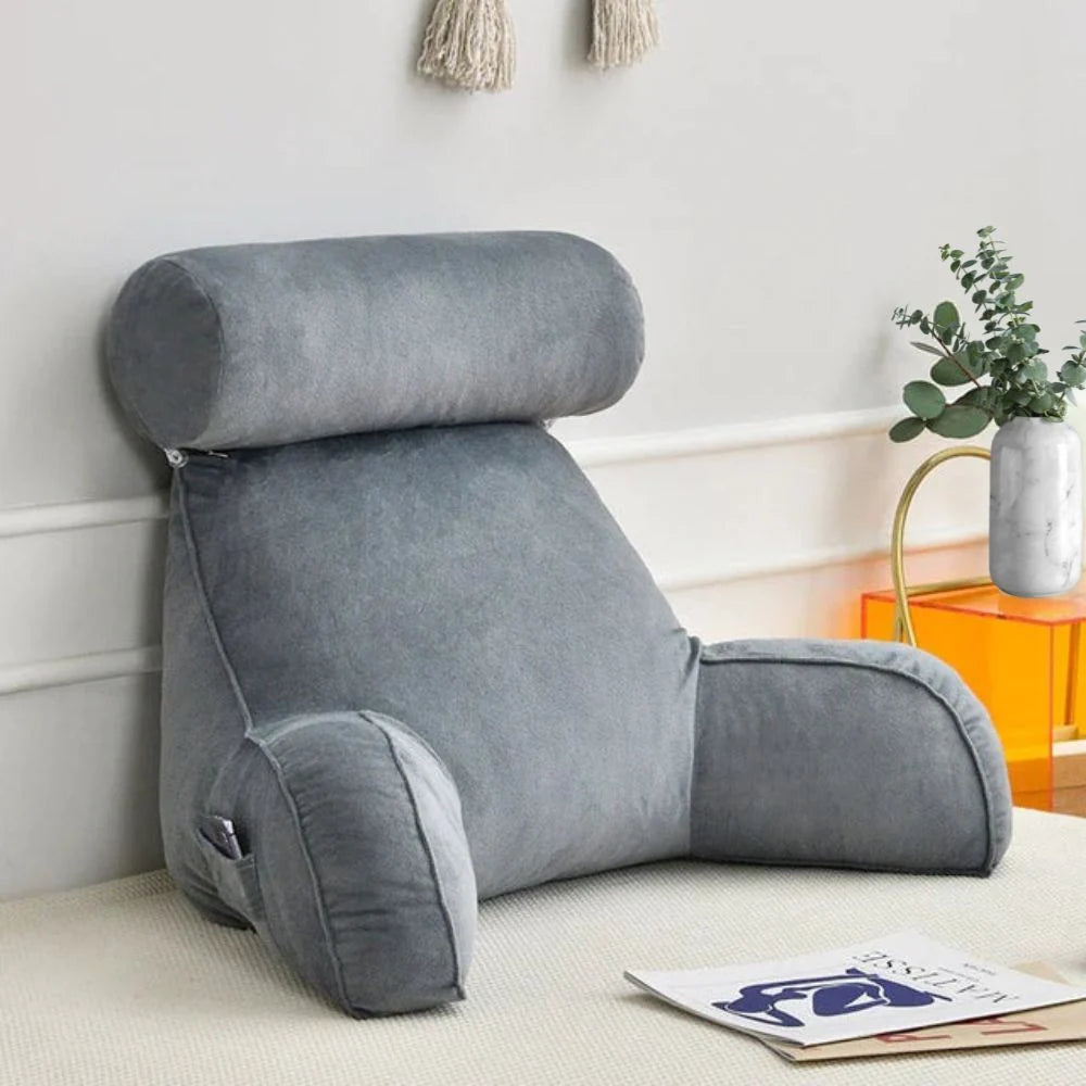 A grey velvet reading pillow with armrests and a detachable headrest, sitting on a bed next to a small marble vase.