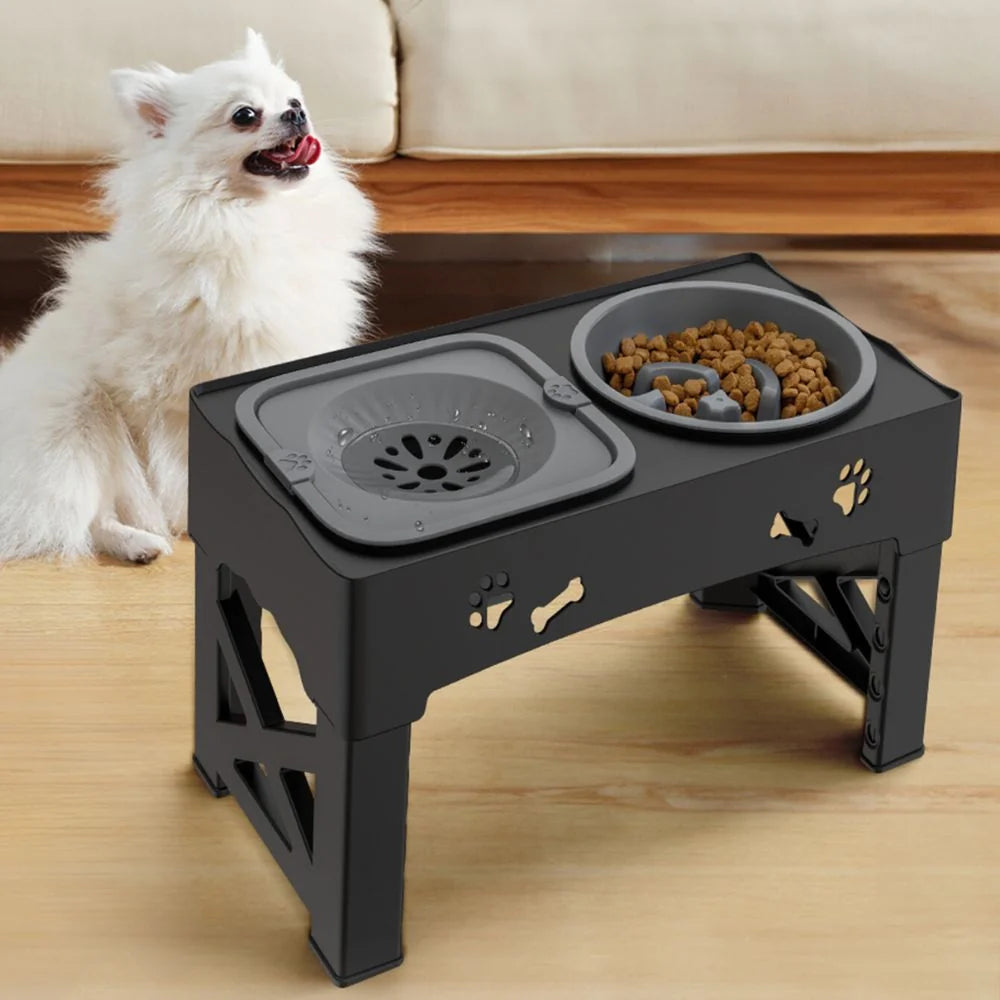 A small white dog sits next to an elevated black pet feeder with a water bowl and a grey slow-feeder bowl for dry food.