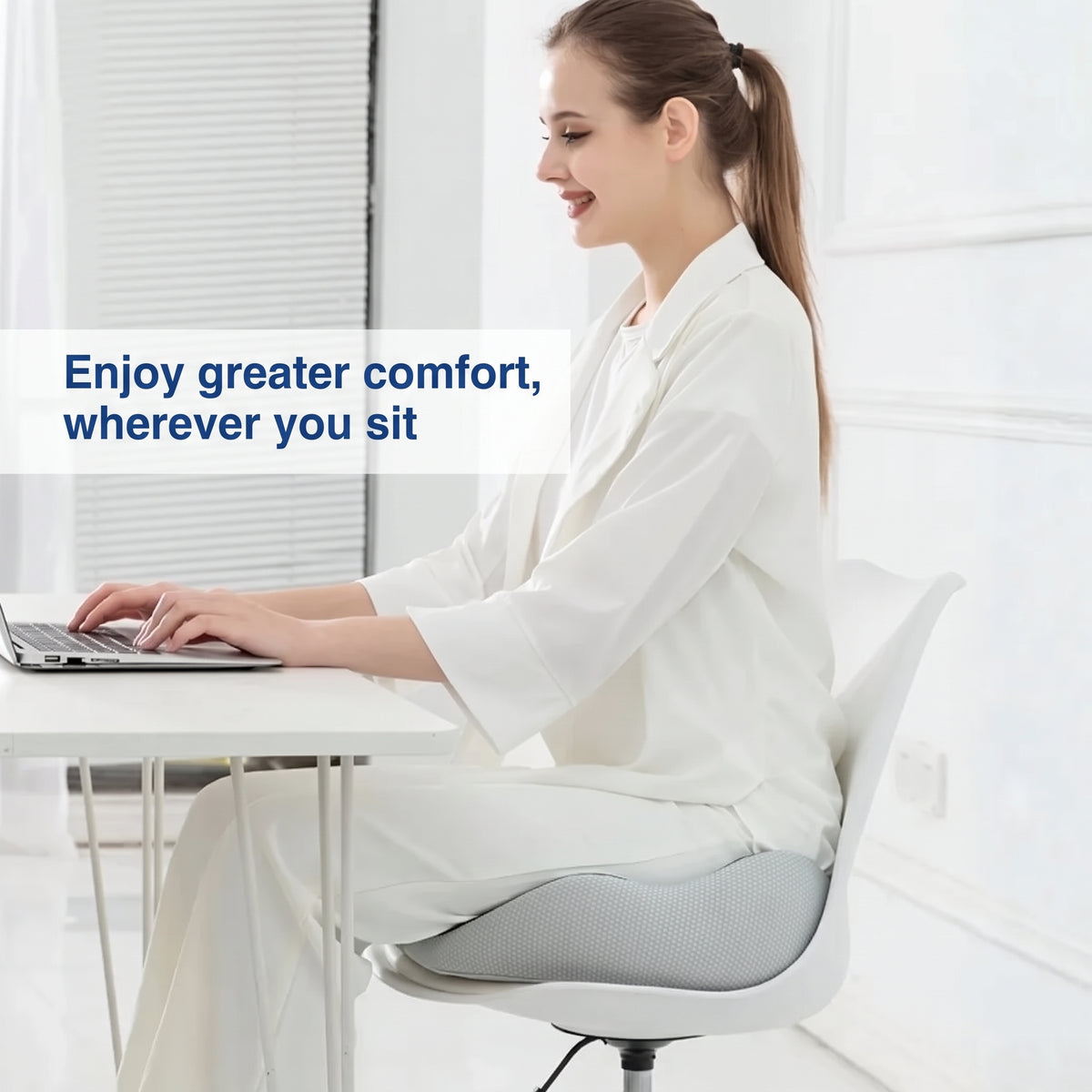 A woman sitting at a desk on a white chair using a grey memory foam seat cushion for added comfort.